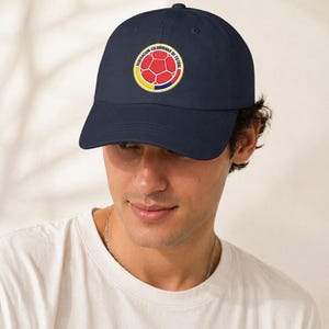 Puede incluir: Gorra de b&eacute;isbol azul marino con un emblema circular con un dise&ntilde;o de bal&oacute;n de f&uacute;tbol rojo, rodeado de anillos amarillos y azules. El texto "FEDERACION COLOMBIANA DE FUTBOL" est&aacute; impreso alrededor del emblema. La gorra tiene un dise&ntilde;o cl&aacute;sico de seis paneles.