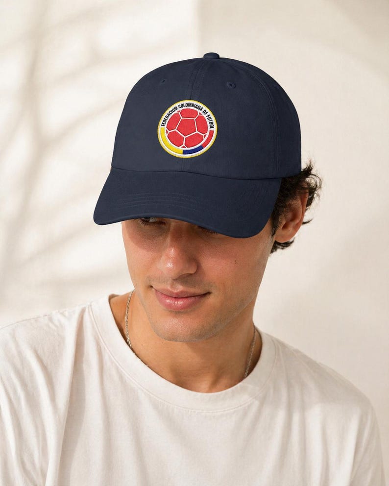 Puede incluir: Gorra de b&eacute;isbol azul marino con un emblema circular con un dise&ntilde;o de bal&oacute;n de f&uacute;tbol rojo, rodeado de anillos amarillos y azules. El texto "FEDERACION COLOMBIANA DE FUTBOL" est&aacute; impreso alrededor del emblema. La gorra tiene un dise&ntilde;o cl&aacute;sico de seis paneles.