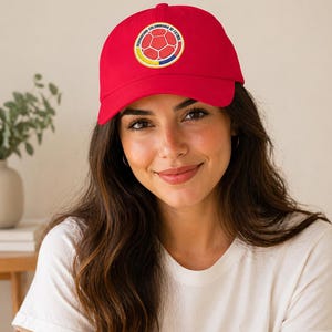 Puede incluir: Gorra de b&eacute;isbol roja con el emblema de la Federaci&oacute;n Colombiana de F&uacute;tbol. La gorra tiene una visera curva y un logotipo circular con un dise&ntilde;o de bal&oacute;n de f&uacute;tbol. La persona lleva una camiseta blanca.