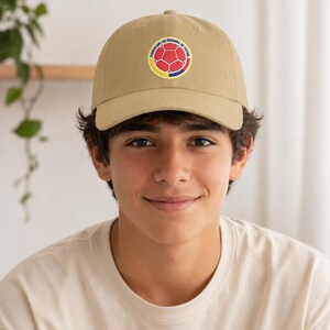 Puede incluir: Gorra de b&eacute;isbol color beige con un emblema circular con un dise&ntilde;o de bal&oacute;n de f&uacute;tbol en rojo, amarillo y azul, con el texto "FEDERACION COLOMBIANA DE FUTBOL". La gorra tiene una visera curva.