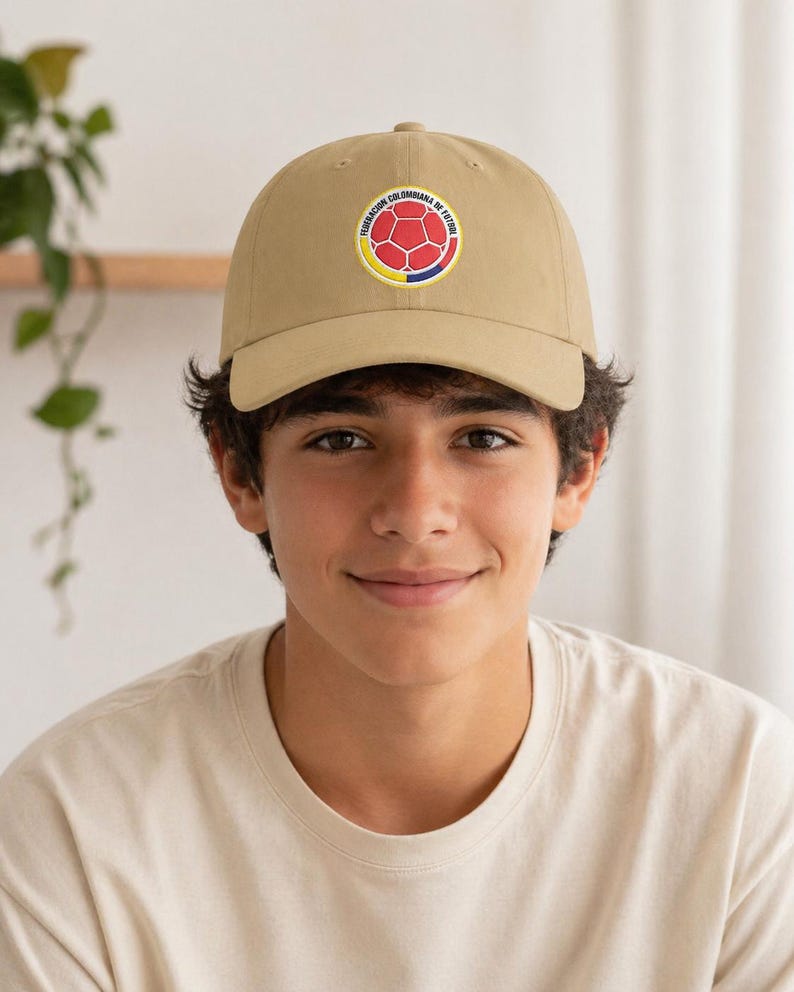 Puede incluir: Gorra de b&eacute;isbol color beige con un emblema circular con un dise&ntilde;o de bal&oacute;n de f&uacute;tbol en rojo, amarillo y azul, con el texto "FEDERACION COLOMBIANA DE FUTBOL". La gorra tiene una visera curva.
