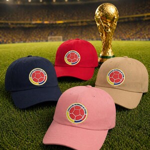 Puede incluir: Cuatro gorras de b&eacute;isbol en azul marino, rojo, beige y rosa, cada una con un emblema circular con un dise&ntilde;o de bal&oacute;n de f&uacute;tbol y texto. Un trofeo dorado est&aacute; en el fondo, sobre un campo verde.