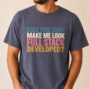May include: A navy blue short-sleeved t-shirt featuring a distressed graphic print with the text "DOES THIS SHIRT MAKE ME LOOK FULL STACK DEVELOPED?" in stacked lines of blue, white, pink, and yellow lettering across the center of the chest.