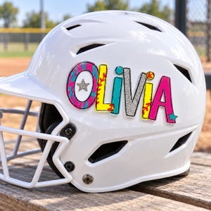 May include: A white batting helmet featuring a custom decal with the name Olivia in colorful, multi-patterned letters. The O is pink and blue with a silver star, the L is yellow with red stitching, the i is teal, the v is silver glitter, the i is yellow, and the A is pink with a teal star.