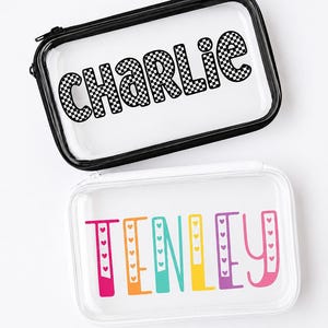 May include: Two rectangular clear zipped pouches. The top pouch has a black zip and features the name CHARLIE in a black and white chequered pattern. The bottom pouch has a white zip and features the name TENLEY in colourful letters.