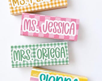 Pode incluir: Quatro carimbos de madeira retangulares com nomes e títulos personalizados. Três apresentam padrões xadrez em amarelo, rosa e verde com texto branco MS. KENDRA, MS. JESSICA e MRS. ORTEGA. Um apresenta um design listrado com SIENNA.