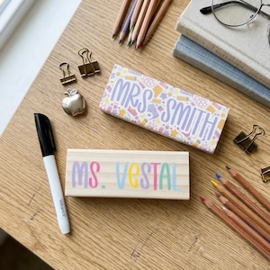 May include: Wooden desk with school supplies, including pencils, books, and a black marker. Two wooden nameplates read "MRS. SMITH" and "MS. VESTAL" in pastel colors. Gold binder clips and a silver apple paperclip are also visible.