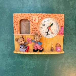 May include: A rectangular wall clock featuring a cracked orange upper section with a calendar block showing the number 2, and a yellow lower section. Two mouse figures are depicted standing on the lower ledge, with a small broom and bucket nearby.