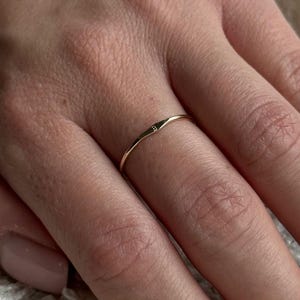 May include: A delicate gold ring with a small, clear gemstone set in the centre. The ring is thin and minimalist, with a slightly textured band. The ring is worn on a finger, showcasing its simple elegance. Jewellery.