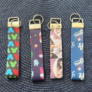 May include: Four keychains with gold-colored hardware. The keychains have different patterns: Mickey Mouse heads, an Among Us character, a woman, and a blue Dodgers baseball team design. The keychains are on a dark blue textured surface.