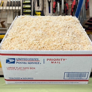 May include: A white cardboard box filled with wood shavings. The box is labeled "United States Postal Service" and "Priority Mail". The box is a "Large Flat Rate Box" with the text "One Rate - Any Weight". The box also has a tracking number: PS00011132700.