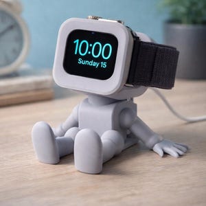 May include: A gray watch charging stand shaped like a seated figure, holding a smartwatch displaying "10:00 Sunday 15". The watch has a black band. The stand is on a wooden surface.
