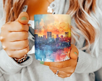 May include: A white ceramic mug with a watercolor cityscape design. The cityscape is painted in shades of blue, orange, yellow, and pink. The mug is being held by a person's hand.