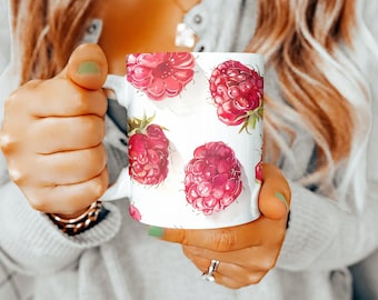 Puede incluir: Taza de cerámica blanca con un patrón de frambuesas rojas. La taza tiene un asa y la está sujetando una persona.