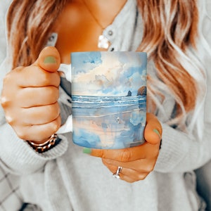 May include: A white ceramic mug with a watercolor design of a beach scene. The design features a blue ocean, white waves, a sandy beach, and a rocky outcropping. The sky is a light blue with white clouds.