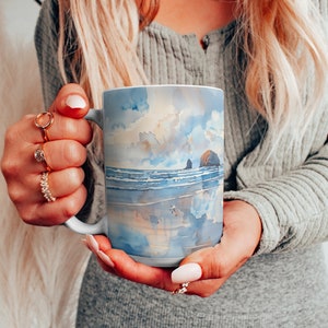 May include: A white ceramic mug with a watercolor design of a beach scene. The design features a blue sky with white clouds, a sandy beach, and a rocky outcropping in the ocean. The mug is being held by a person's hands.