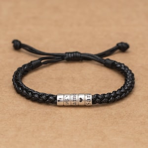 Può includere: Bracciale in pelle intrecciata nera con perline d'argento. Le perline sono incise con nomi e simboli, tra cui un cuore. Il bracciale è regolabile e ha una chiusura annodata. Un accessorio personalizzato.