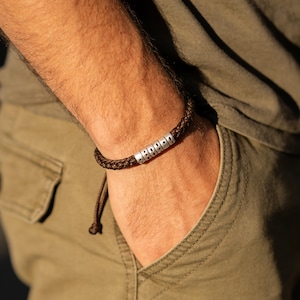 May include: A brown braided bracelet with silver beads. The bracelet is worn on a person's wrist. The person is wearing an olive green shirt and khaki pants. The bracelet has a simple, classic design.