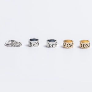 Puede incluir: Una colección de joyas que incluye dos anillos de plata pequeños con superficies texturizadas, seguidos de tres anillos de banda más anchos. Las bandas presentan texto grabado que dice MIKEY, Tom y JACK, mientras que el último anillo dorado muestra el número 1923.