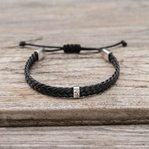 May include: A black braided cord bracelet featuring a silver-toned metal bead engraved with a bicycle icon. The bracelet includes two silver-toned end caps and an adjustable sliding knot closure mechanism made from the same black cord material.