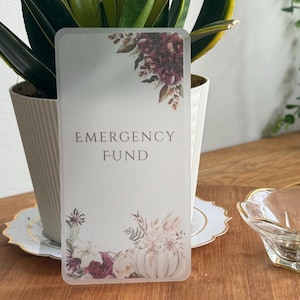 May include: A rectangular card with the words "EMERGENCY FUND" in the centre. The card has floral and pumpkin designs in the corners. The text "Velvet Pumpkin Reserve" is at the bottom. The card is on a wooden surface.