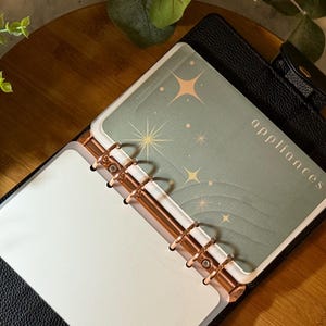 May include: A black leather-textured ring binder planner featuring rose gold metal rings. Inside, a sage green divider tab is printed with the word appliances and decorated with gold star icons. The planner is open, revealing blank white pages.