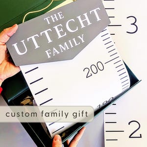 May include: A custom family gift set featuring a gray sign with "THE UTTECHT FAMILY" in white lettering. The set includes a green box and a height chart with black markings and numbers. The height chart measures up to 3 meters.