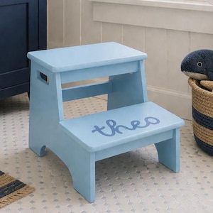 May include: A light blue wooden two-step stool featuring the name theo written in a darker blue cursive script on the bottom step. The stool has cut-out handles on the sides of the top step and a smooth finish throughout the entire structure.