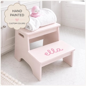May include: A light pink wooden two-step stool featuring the name "ella" in cursive pink lettering on the bottom step. A white folded towel and a small pink round figurine with a face rest on the top step of the stool.