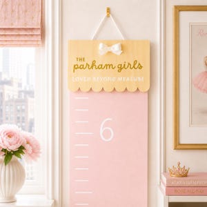 May include: A light pink wooden growth chart hanging by a white cord. The top features a scalloped edge, a white bow, and the gold text "THE parham girls LOVED BEYOND MEASURE". The chart includes white measurement markings and the number 6.