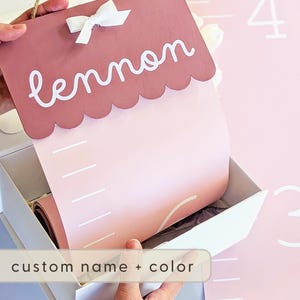 May include: A custom height chart in a white box. The chart is pink and has a scalloped top with the name "lennon" in white script. A white bow is attached. The chart is designed to measure height and has white markings. The text "custom name + color" is visible.