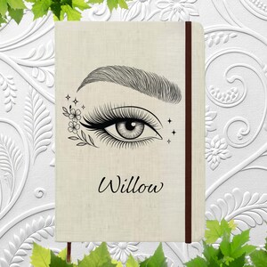 May include: A beige journal with a detailed eye and eyebrow illustration, floral accents, and the name "Willow" in script. The journal has a brown elastic closure.