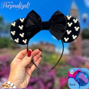 May include: Black velvet mouse ear headband with a large bow and white Mickey Mouse head accents. The text "Can be Personalized!" is in the upper left corner. The headband is held by a person.