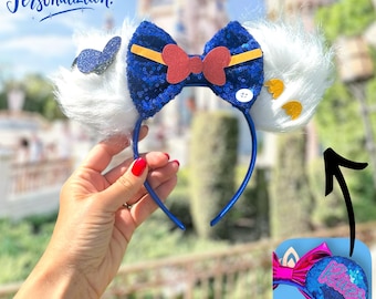 Puede incluir: Una diadema azul con un lazo de lentejuelas y orejas blancas y esponjosas. El lazo tiene un centro rojo y detalles amarillos. Una mariposa azul y detalles amarillos están en las orejas. El texto "Free Personalization!" está en la esquina superior izquierda.