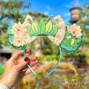 May include: A headband with a beige sequined bow, a green glitter crown, and two side accents with pink flowers and frog illustrations. The text "Can be Personalized!" is visible.