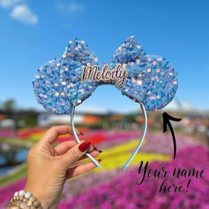 May include: Blue sequin Minnie Mouse ears headband with a bow and the name "Melody" embroidered on the bow.  The headband is held in a hand with red nail polish.