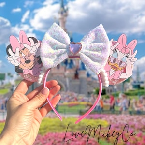 May include: Pink Minnie Mouse and Daisy Duck ear headband with a large sequined bow and a heart-shaped gem. The headband has a pink frame and white pearl accents. The text "Love Mickey Co" is at the bottom.