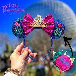 May include: A blue headband with sequined ears featuring a pink flower design and a pink bow. A gold crown sits atop the bow. The text "Free Personalization!" is visible in the upper left corner.