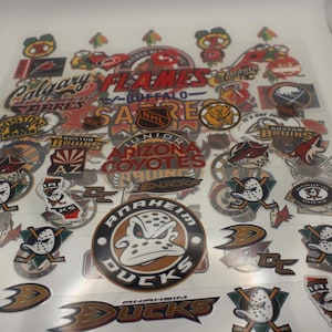 Könnte beinhalten: Eine Sammlung von Hockey-Team-Logo-Aufklebern, darunter die Anaheim Ducks und Boston Bruins. Die Aufkleber zeigen Teamlogos und Maskottchen in verschiedenen Farben und Designs, ideal für Sportfans.