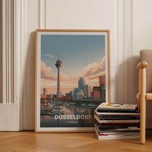 Op de afbeelding: Een ingelijste reisposter met een gestileerde illustratie van de skyline van Düsseldorf bij zonsondergang. De afbeelding toont de Rheinturm, moderne gebouwen en boten op het water, met de tekst DÜSSELDORF GERMANY aan de onderkant.