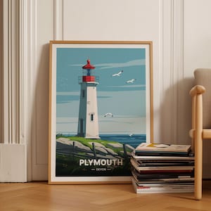 Puede incluir: Una impresión enmarcada de un faro con techo rojo y paredes blancas. El faro está en una costa rocosa con hierba verde y un cielo azul con pájaros blancos volando por encima. La impresión tiene el texto "PLYMOUTH - DEVON" en la parte inferior.
