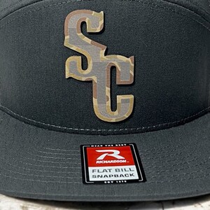 May include: A gray and tan camouflage flat bill snapback hat with a large "SC" logo on the front. The hat is made by Richardson.