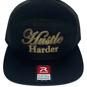 May include: Black and gold flat bill trucker hat with the words "Hustle Harder" embroidered on the front.