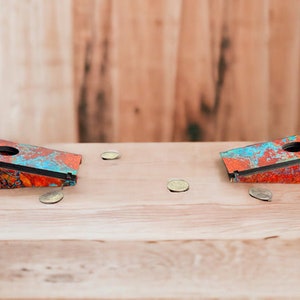 May include: Two wooden bean bag toss game boards with a colorful abstract design in shades of orange, blue, and black. The boards are sitting on a wooden surface with four silver coins scattered around them.