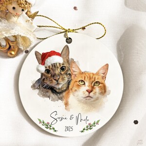 Puede incluir: Adorno navideño redondo de cerámica blanca con retratos de acuarela de dos gatos. Un gato lleva un gorro de Papá Noel. El adorno está personalizado con los nombres "Suzie & Milo" y el año "2025", con vegetación decorativa.