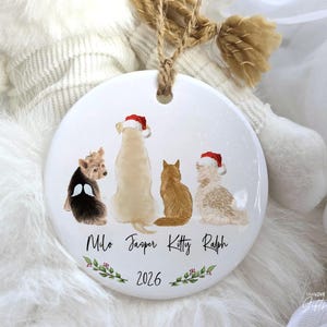 May include: A round white ceramic ornament hanging from a jute twine loop. The front features illustrations of four pets, two wearing Santa hats, with the names Milo, Jasper, Kitty, and Ralph printed below, followed by the year 2026 and a holly sprig.
