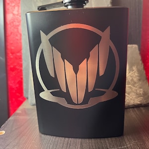 Puede incluir: Petaca negra de acero inoxidable con un diseño de emblema plateado. La petaca tiene una tapa de rosca negra y una tapa con bisagras. El emblema es un diseño simétrico estilizado dentro de un círculo.