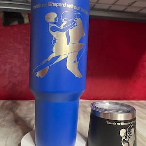 Puede incluir: Dos vasos de acero inoxidable azules con un diseño plateado de dos figuras bailando. Los vasos tienen el texto "There's no Shepard without Vakarian".