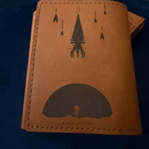 Op de afbeelding: Bruin leren portemonnee met een zwart grafisch ontwerp uit de Mass Effect-videogameserie. Het ontwerp bevat een silhouet van een figuur, een groot wezen en de tekst "MASS EFFECT".