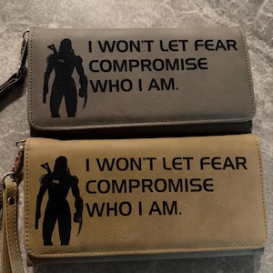 Op de afbeelding: Twee leren portemonnees, één grijze en één bruin, elk met een zwart silhouet van een figuur en de tekst "I WON'T LET FEAR COMPROMISE WHO I AM." Beide portemonnees hebben een polsband en een ritssluiting.